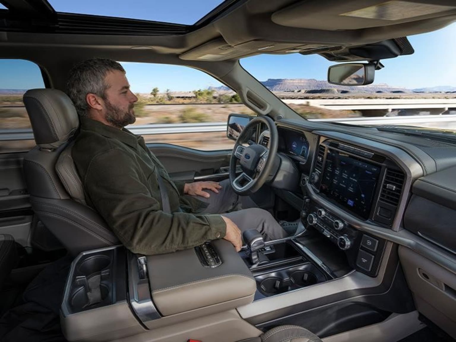 A man with a beard sitting in the driver's seat of a large vehicle, surrounded by a desert landscape in the background.