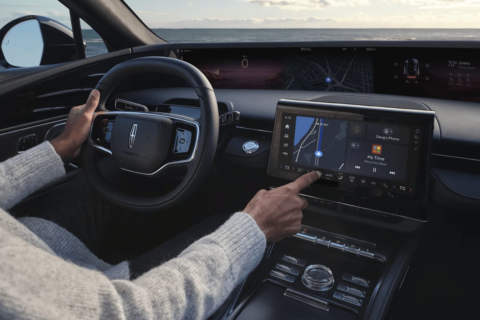 The image shows the interior of a modern vehicle, with the driver's hands on the steering wheel and dashboard controls. The background outside the windshield depicts a scenic landscape with a cloudy sky.