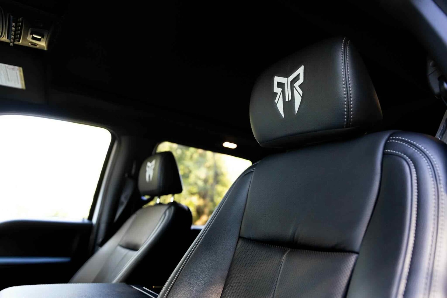 Black leather seats with a white logo in the foreground, blurred outdoor scene with trees visible through the windows in the background.