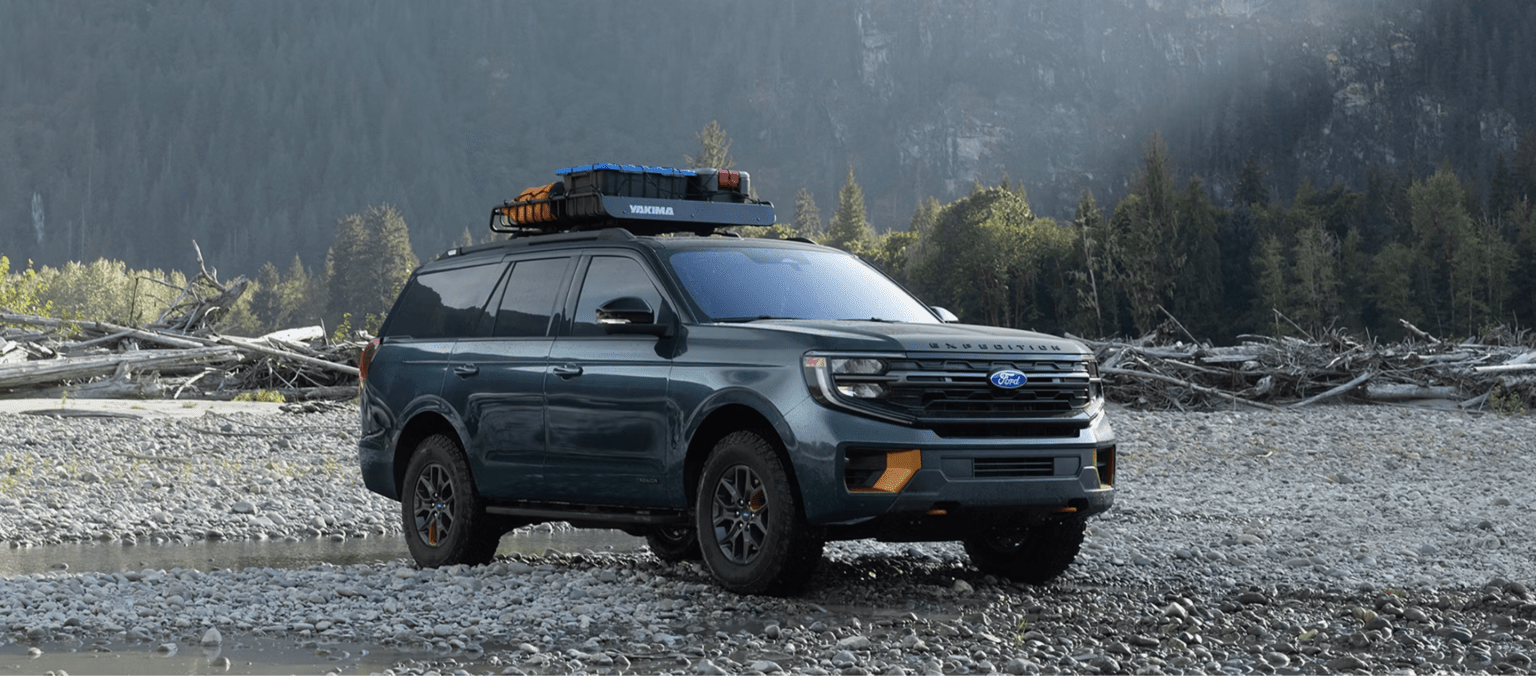 Dark green SUV with roof rack parked on rocky riverbed in forest/mountain setting
