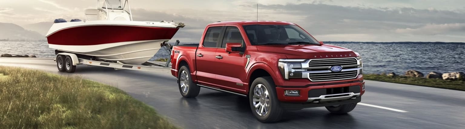 A red pickup truck towing a boat on a trailer, with a scenic coastal landscape in the background featuring a cloudy sky and water.