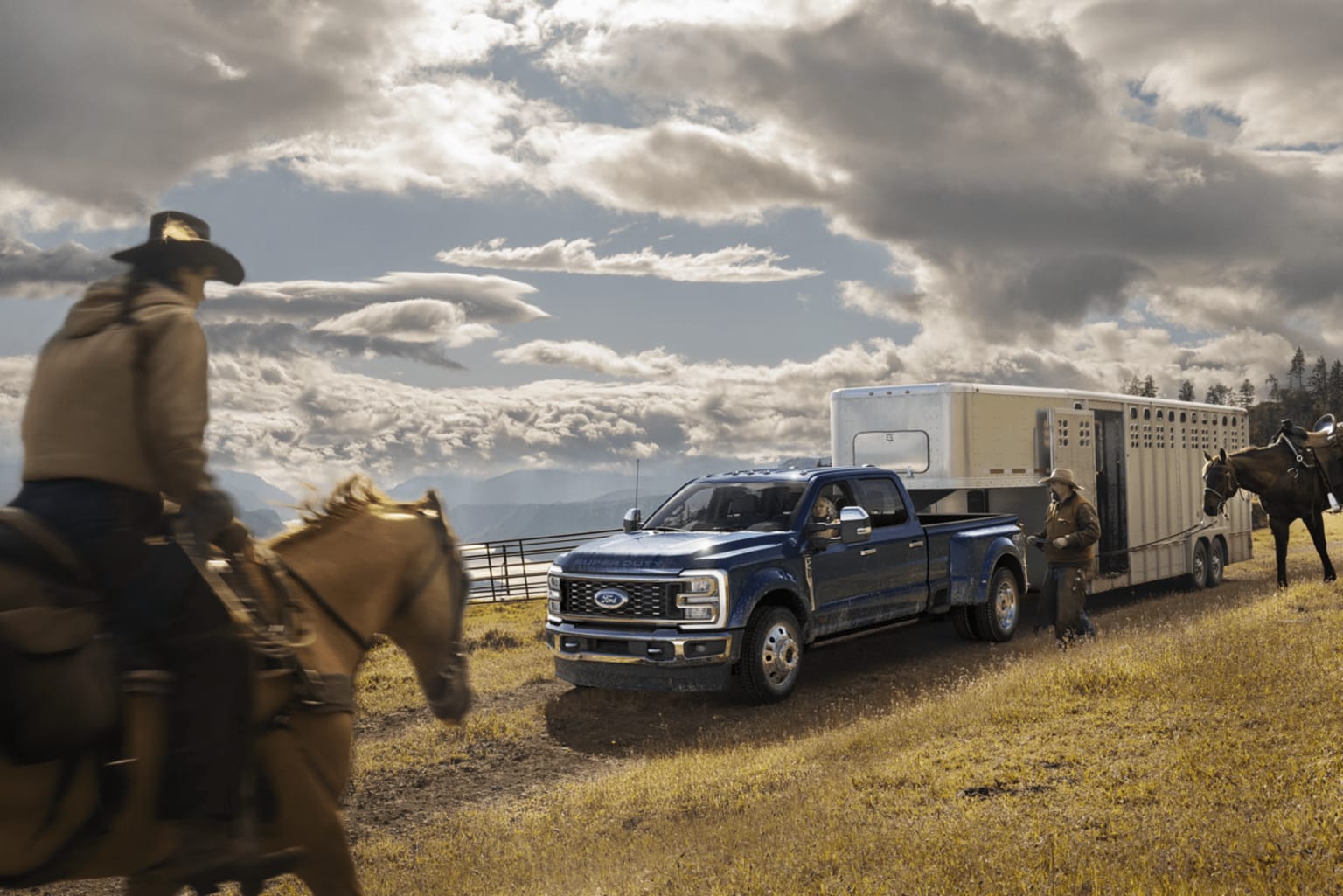 A pickup truck towing a trailer is parked in a grassy field, with horses visible in the background and dramatic cloudy skies overhead.