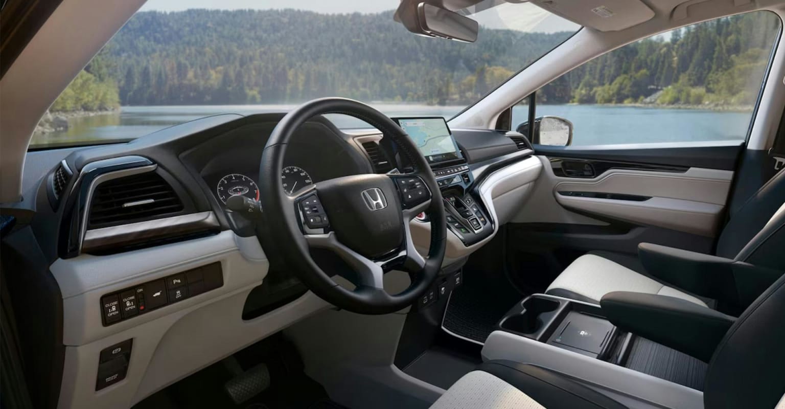 Interior of a modern car with a steering wheel and dashboard, with a serene lake and forested hills in the background.