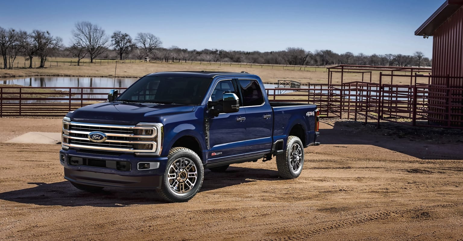 A blue pickup truck parked in a rural setting with a pond, fences, and trees visible in the background.