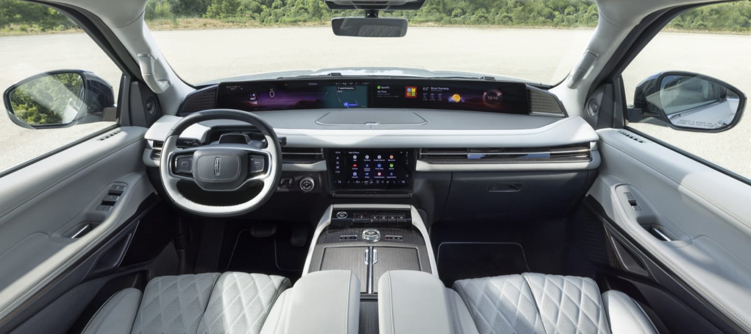 The image shows the interior of a modern vehicle with a sleek dashboard, a large central display, and various controls and indicators. The background outside the vehicle appears to be a natural setting, with trees and greenery visible through the windshield.