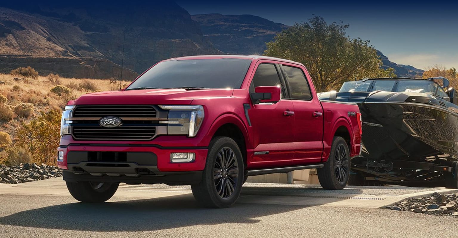 A bright red pickup truck stands prominently in the foreground, surrounded by a rugged, mountainous landscape in the background.