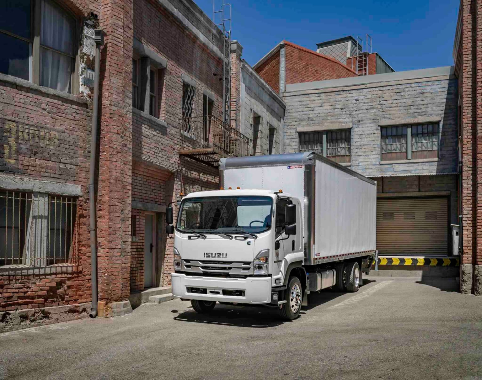 A white box truck is parked in a narrow alleyway between brick buildings, with a large garage door visible in the background.