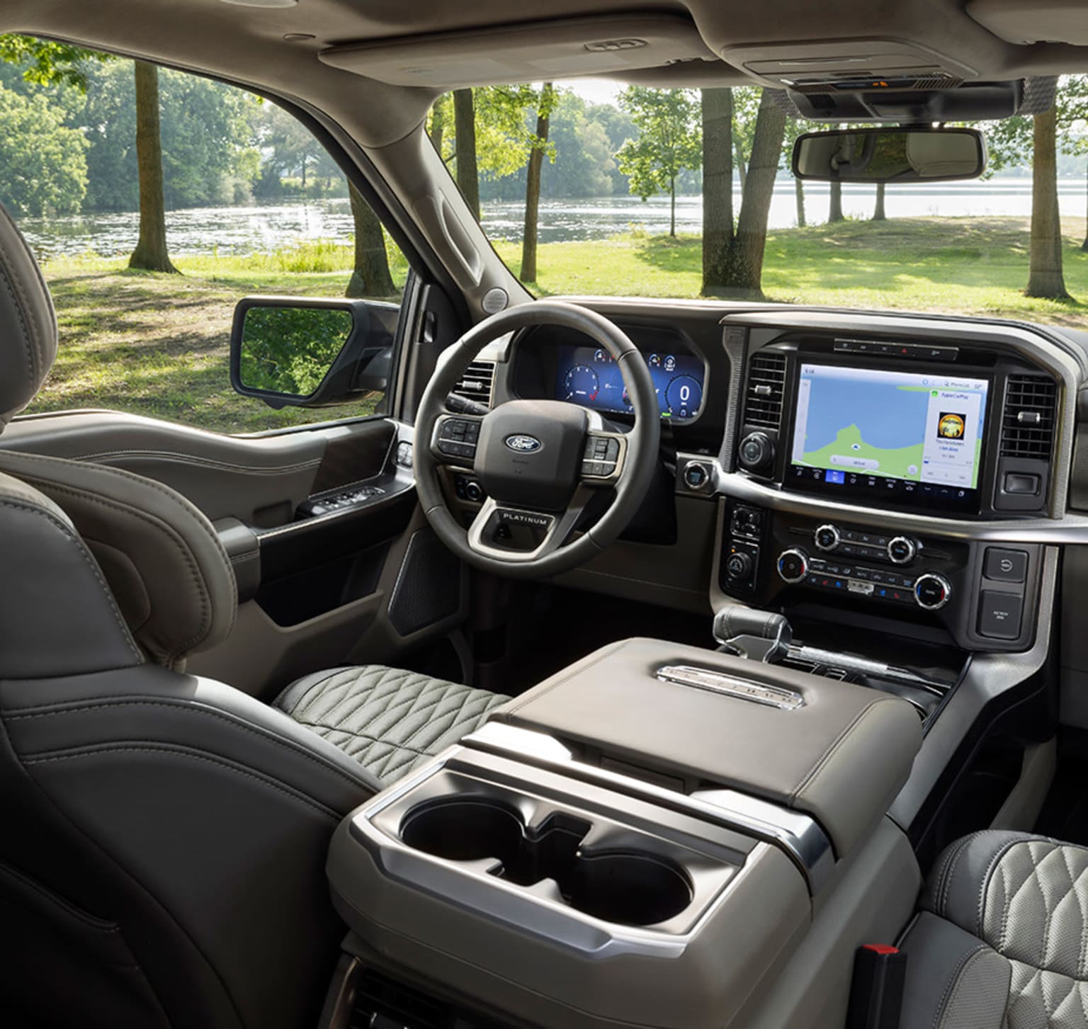Interior of a modern vehicle with large touchscreen display, steering wheel, controls, and features, set against a lush green park or wooded area.