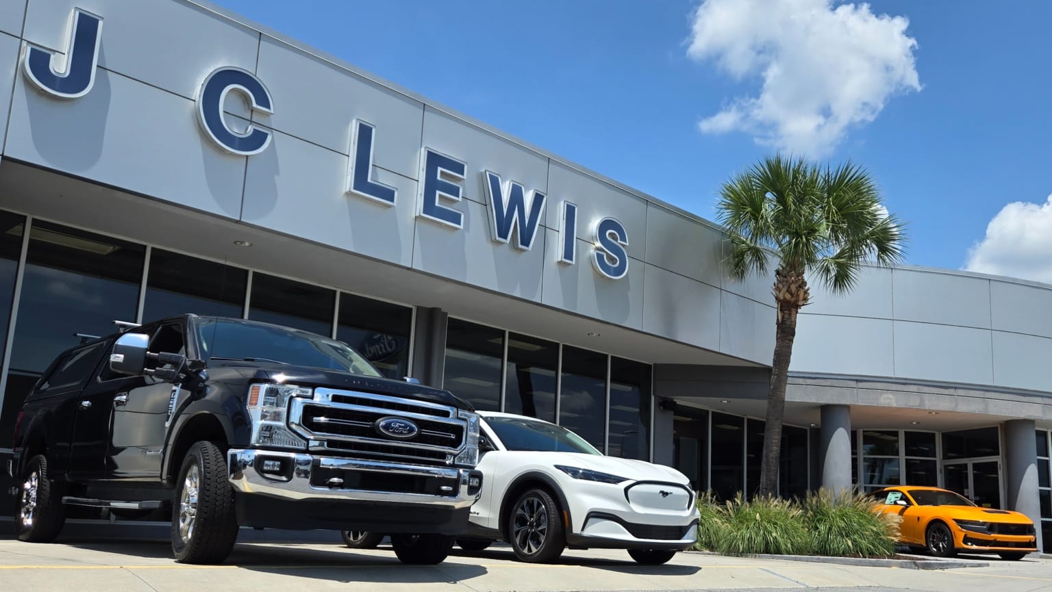 A black truck and a white electric vehicle are parked in front of a dealership, with a yellow sports car visible to the right and a palm tree against a cloudy blue sky in the background.