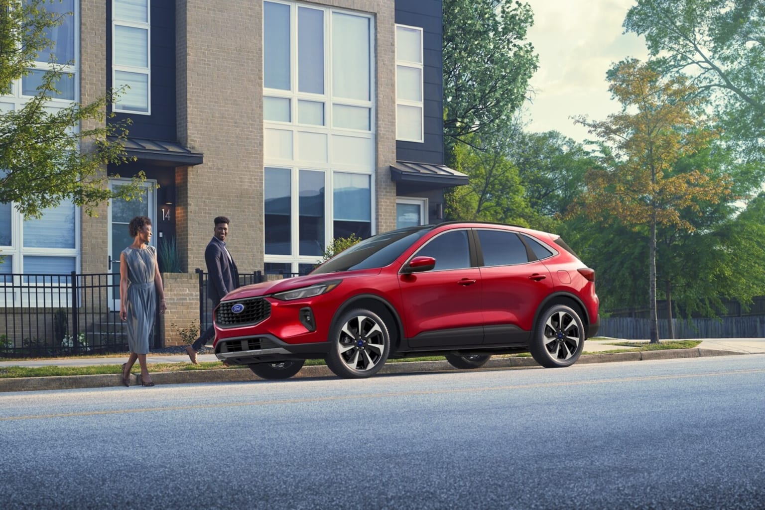A red SUV parked in front of a modern multi-story building, surrounded by trees and a paved road.