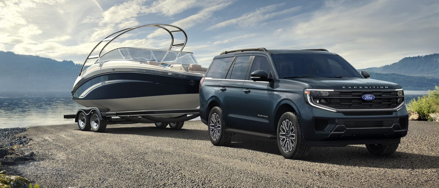 A dark blue SUV tows a boat on a trailer along a gravel path next to a calm lake with mountains in the background.