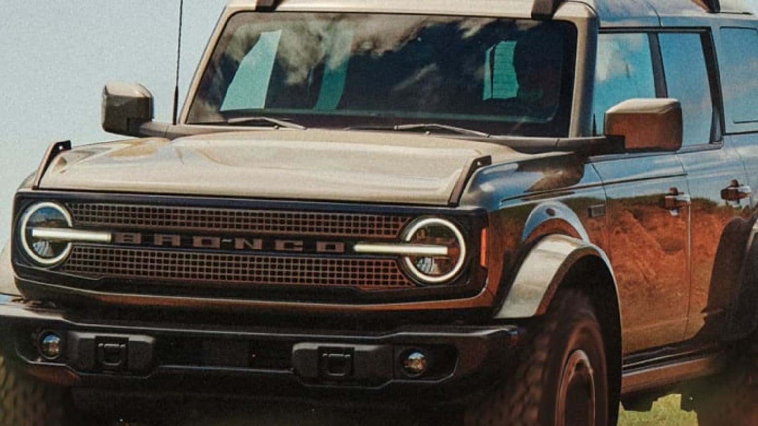 A tan SUV with a black grille and round headlights is parked on a dirt road with a blurred background of trees and sky.