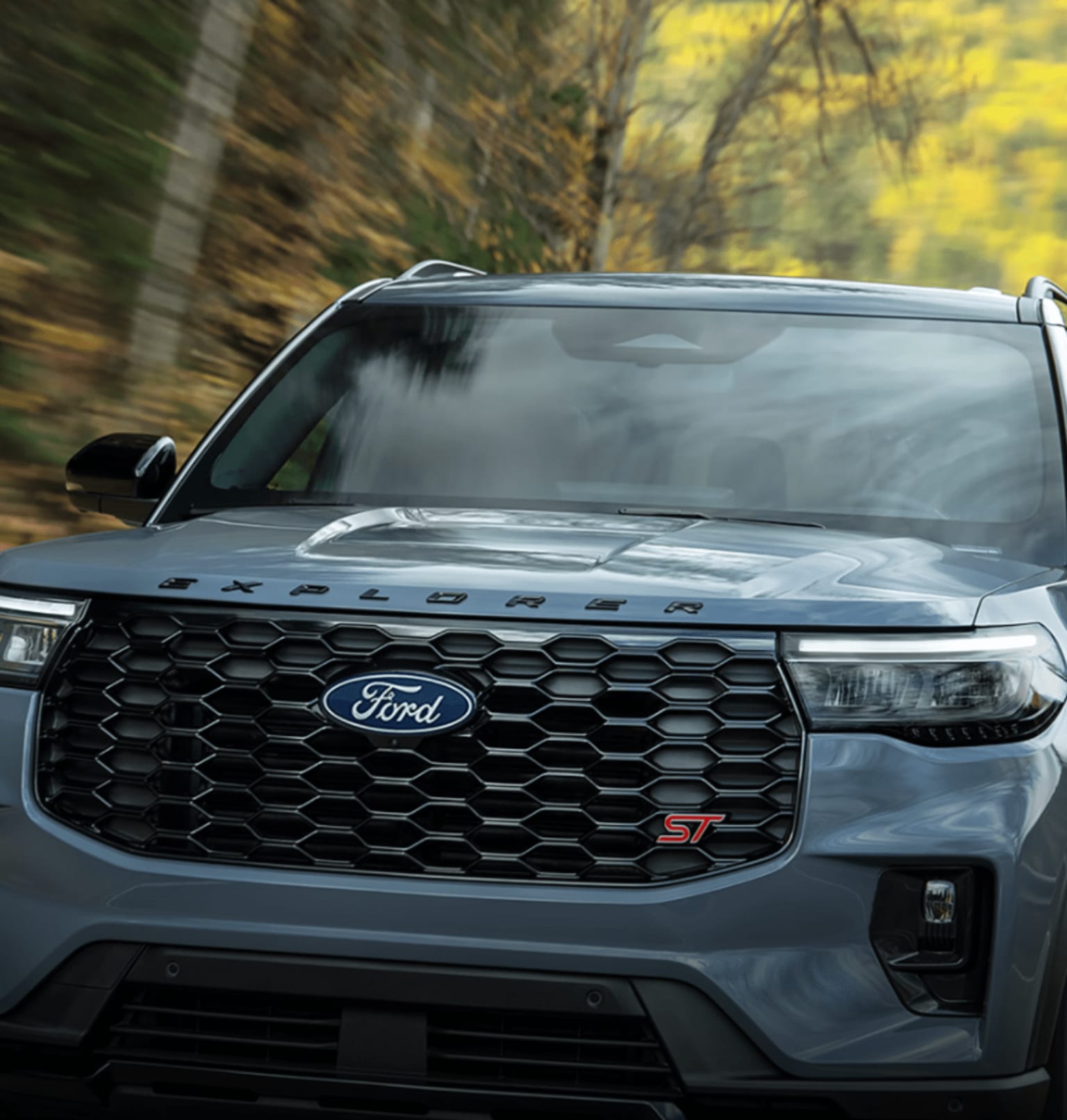 A grey SUV is parked in front of autumn trees.