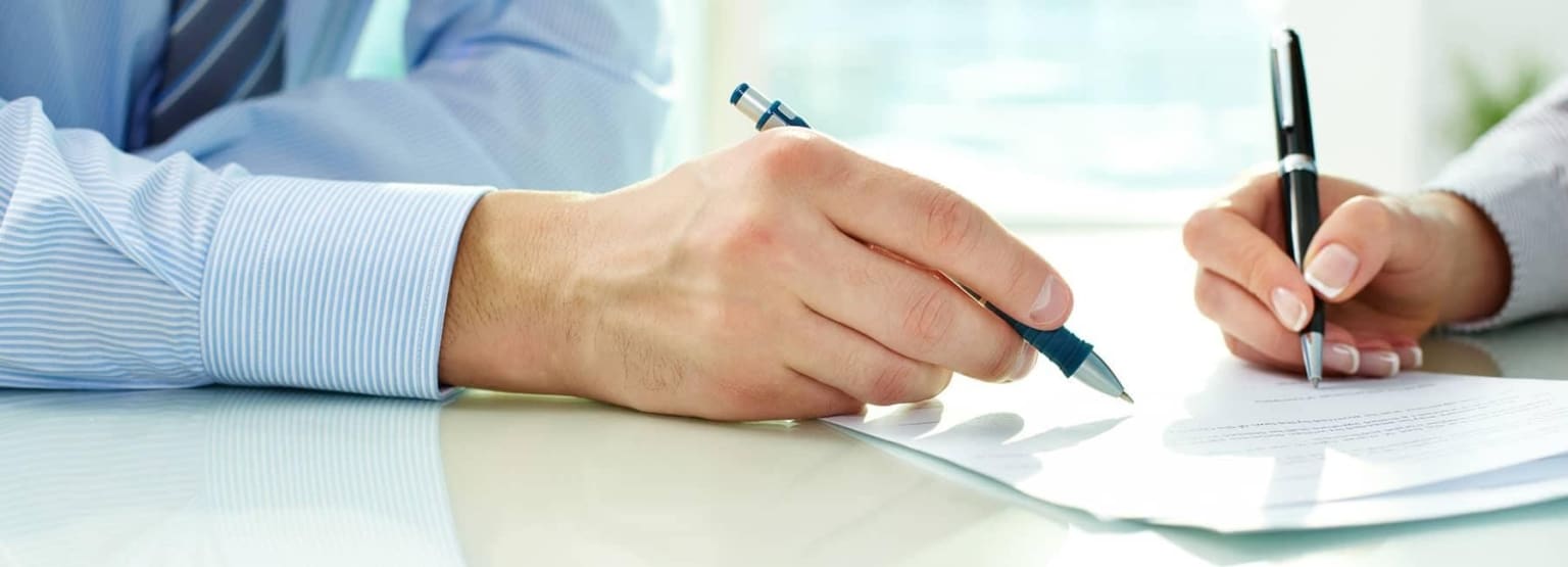 Two individuals in business attire shaking hands over a document on a desk with a pen in one hand.