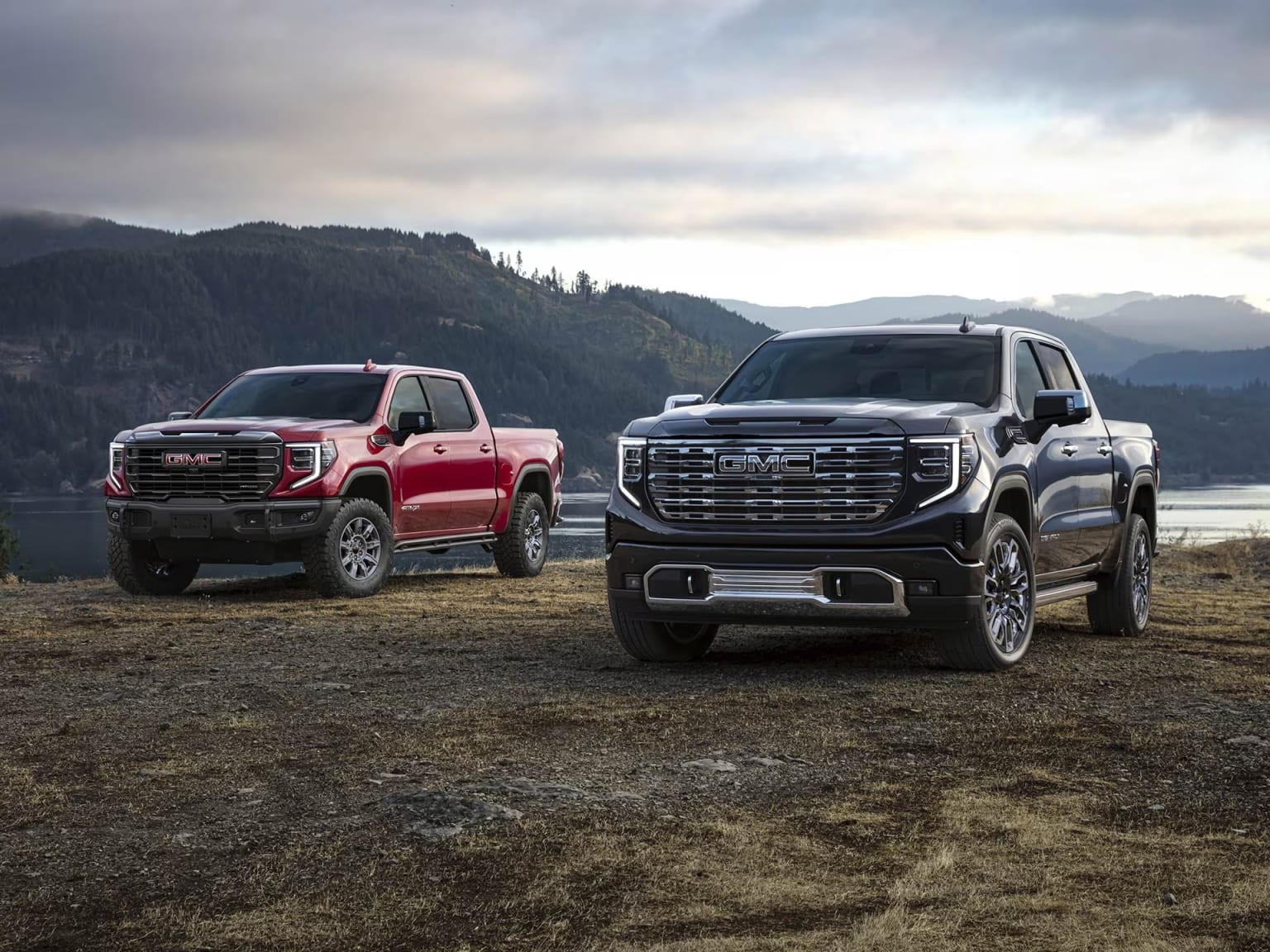 Two pickup trucks parked on grass with a mountainous background under a cloudy sky.