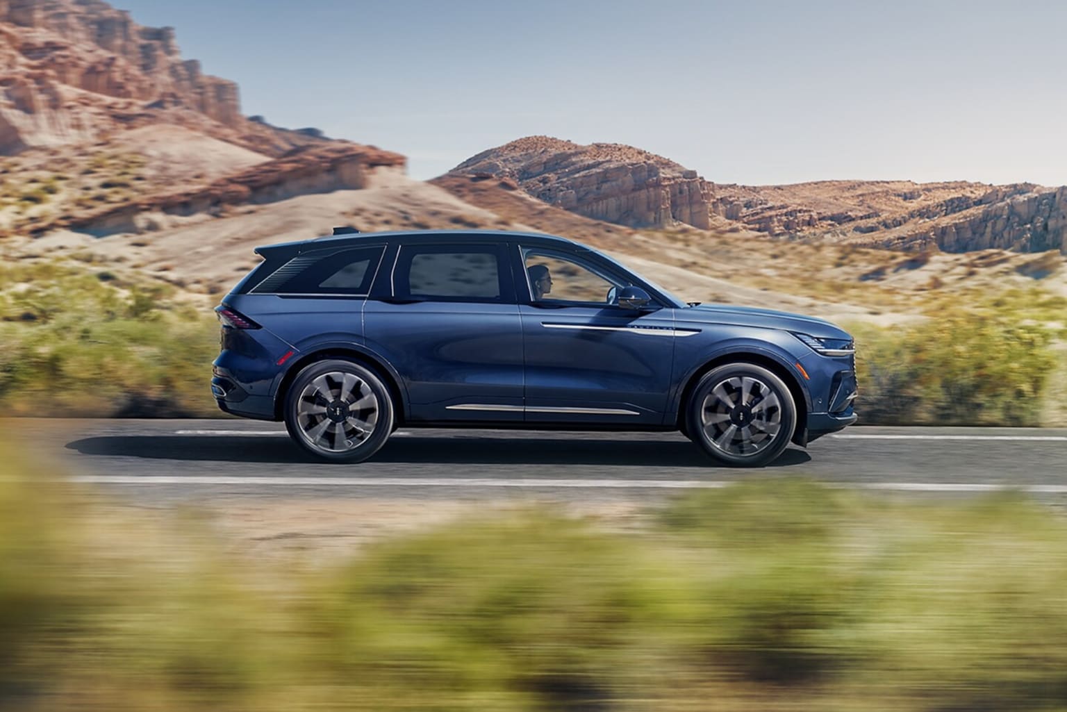 A sleek, dark-colored SUV driving through a rugged, mountainous desert landscape with towering rock formations in the background.