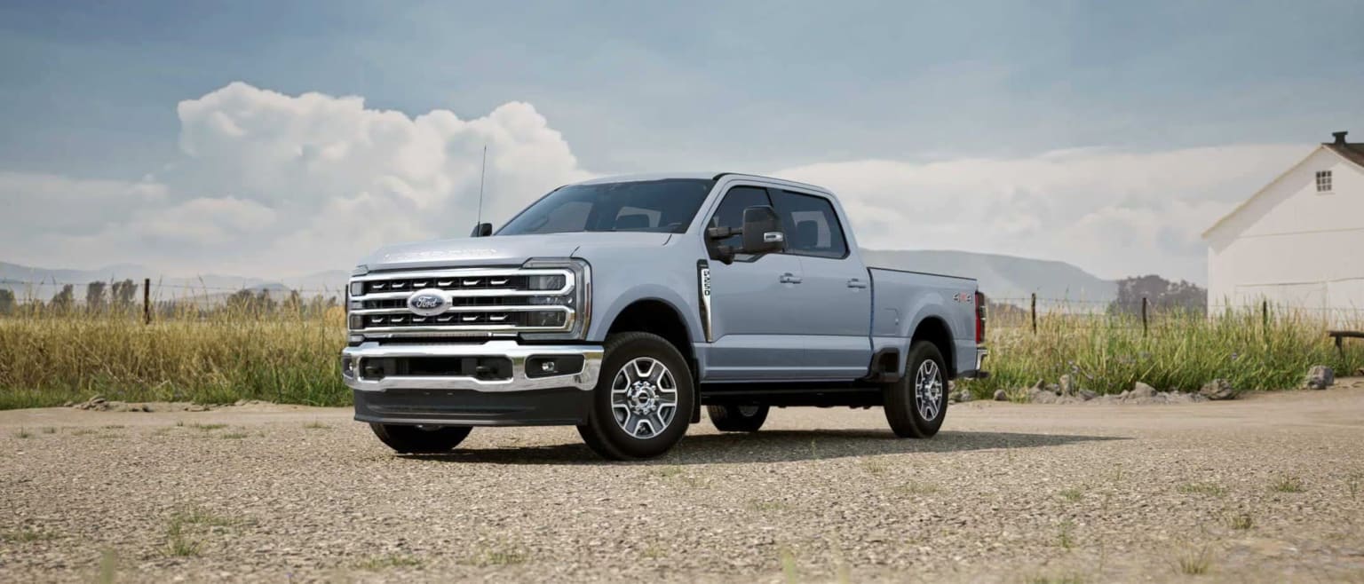 A large, silver pickup truck on a dirt road in a grassy field with cloudy sky.