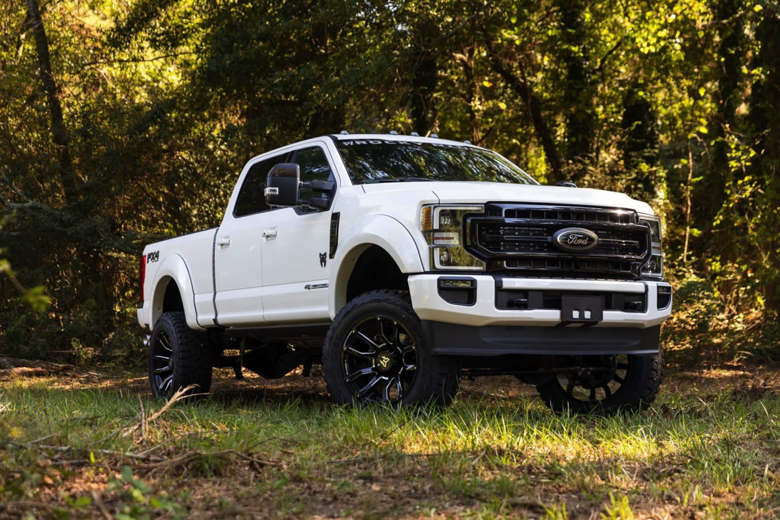 A white pickup truck stands prominently in a lush, forested setting, its large tires and lifted suspension suggesting its off-road capabilities.