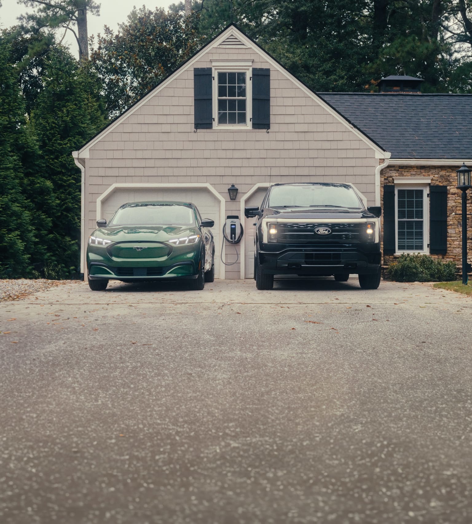 A two-car garage with a gray exterior and black shutters sits in a wooded area, with a green vehicle and a black truck parked in the driveway.