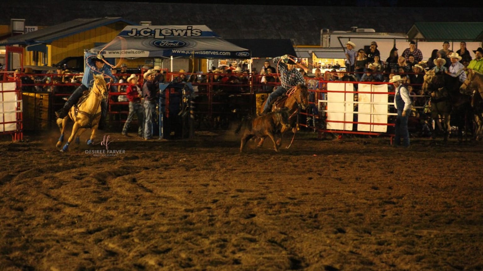 a couple of men riding on the backs of horses in a dirt arena at night time with a crowd watching