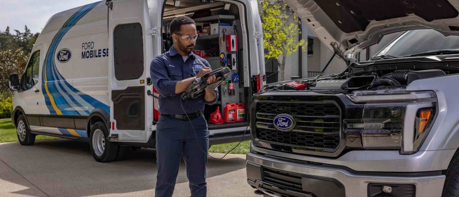 A mechanic in a mobile service van works on a truck with its hood open in front of a suburban house.