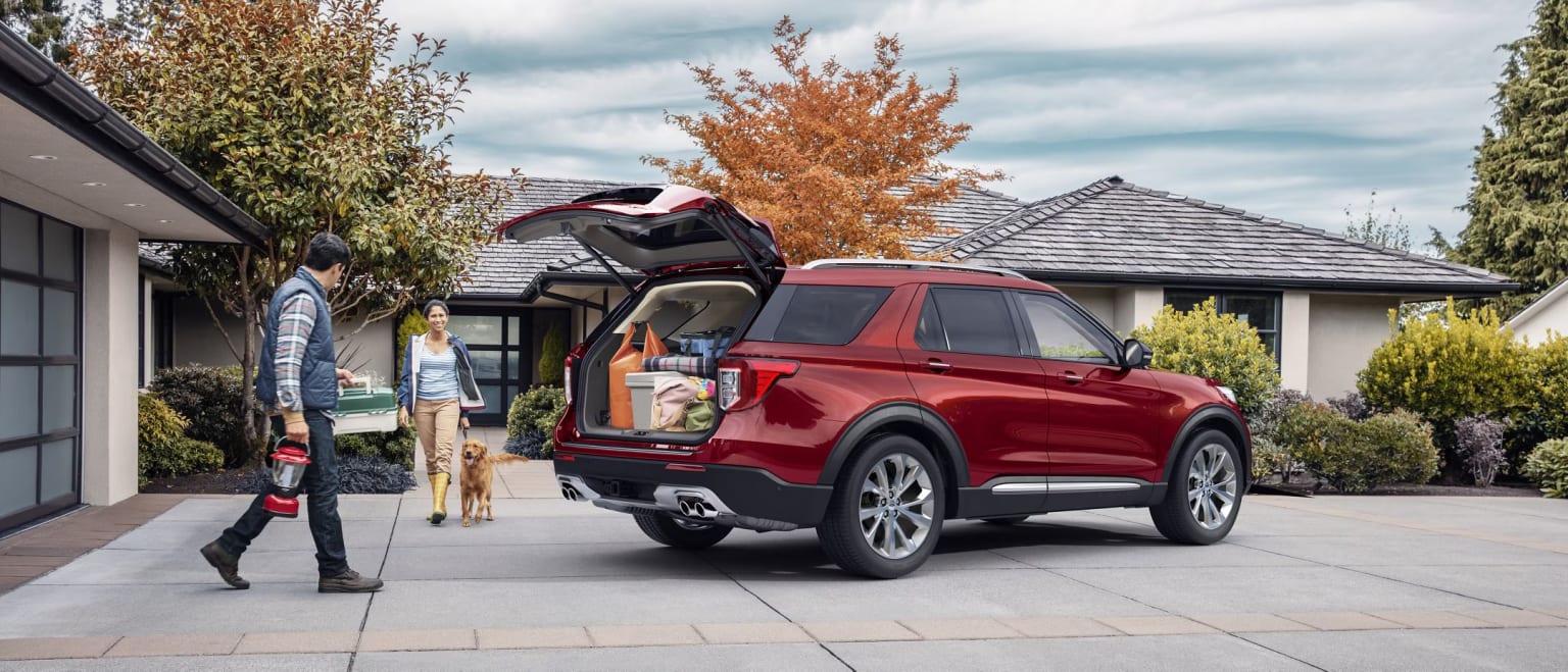 A red SUV with an open rear hatch is parked in front of a residential home with a tiled roof, surrounded by autumn foliage.