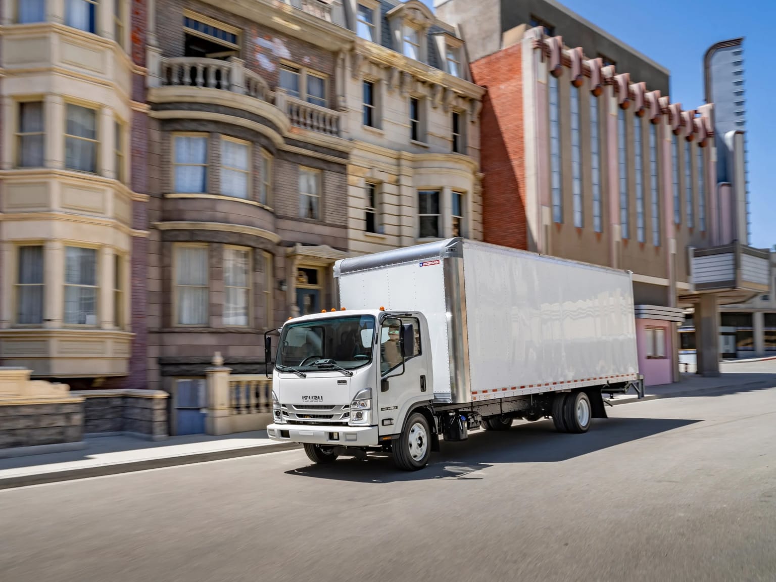 A white delivery truck driving down a city street with ornate, historic-looking buildings in the background.