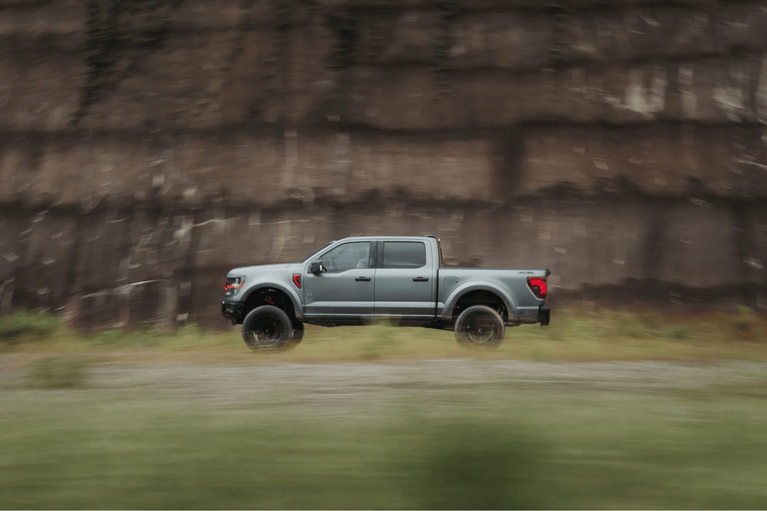 A large, lifted pickup truck off-roading through a muddy, grassy field, with a blurred background of a concrete wall.