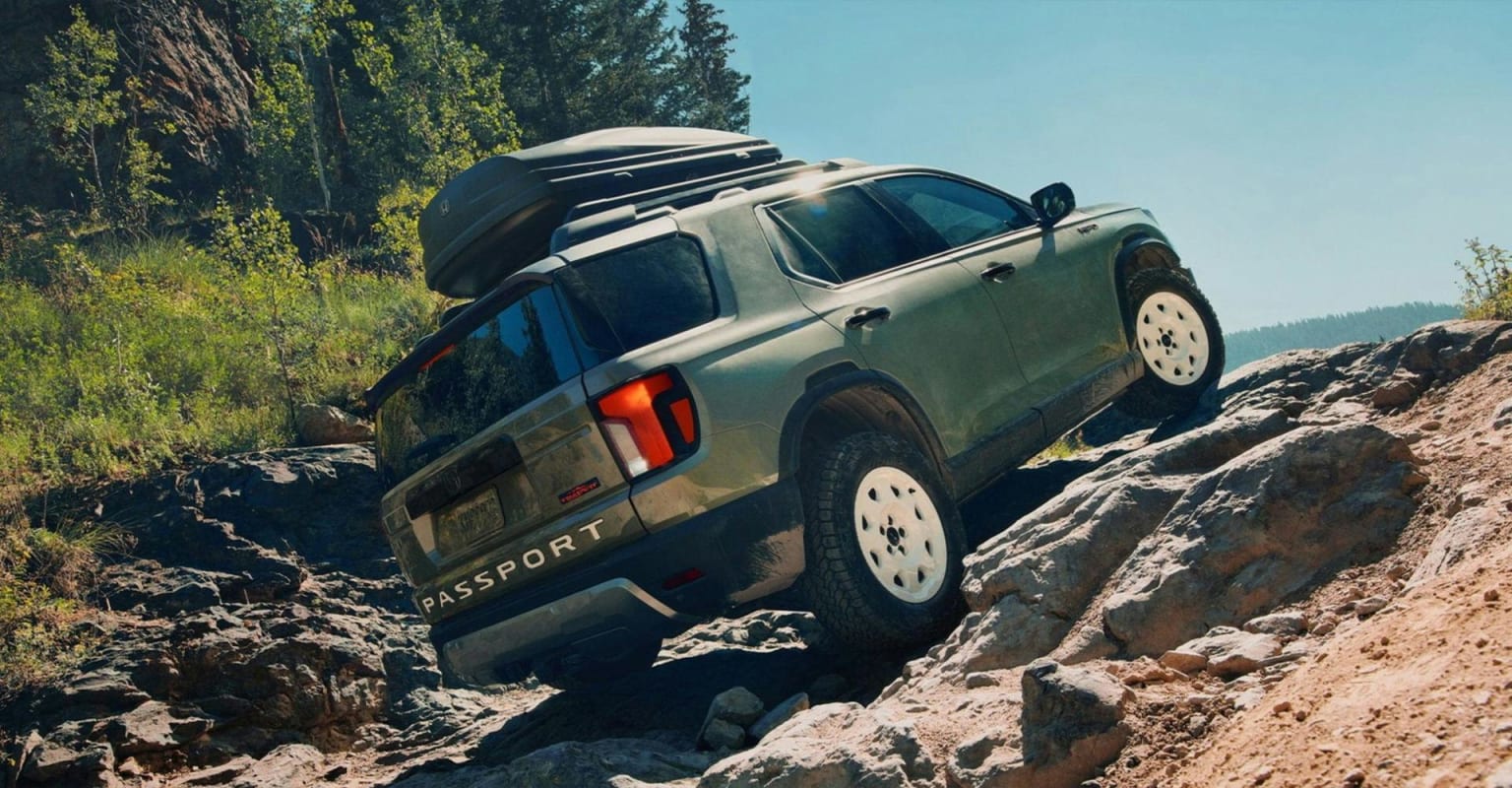 A green SUV with a roof rack navigating a rocky, uphill trail with a wooded hillside in the background.