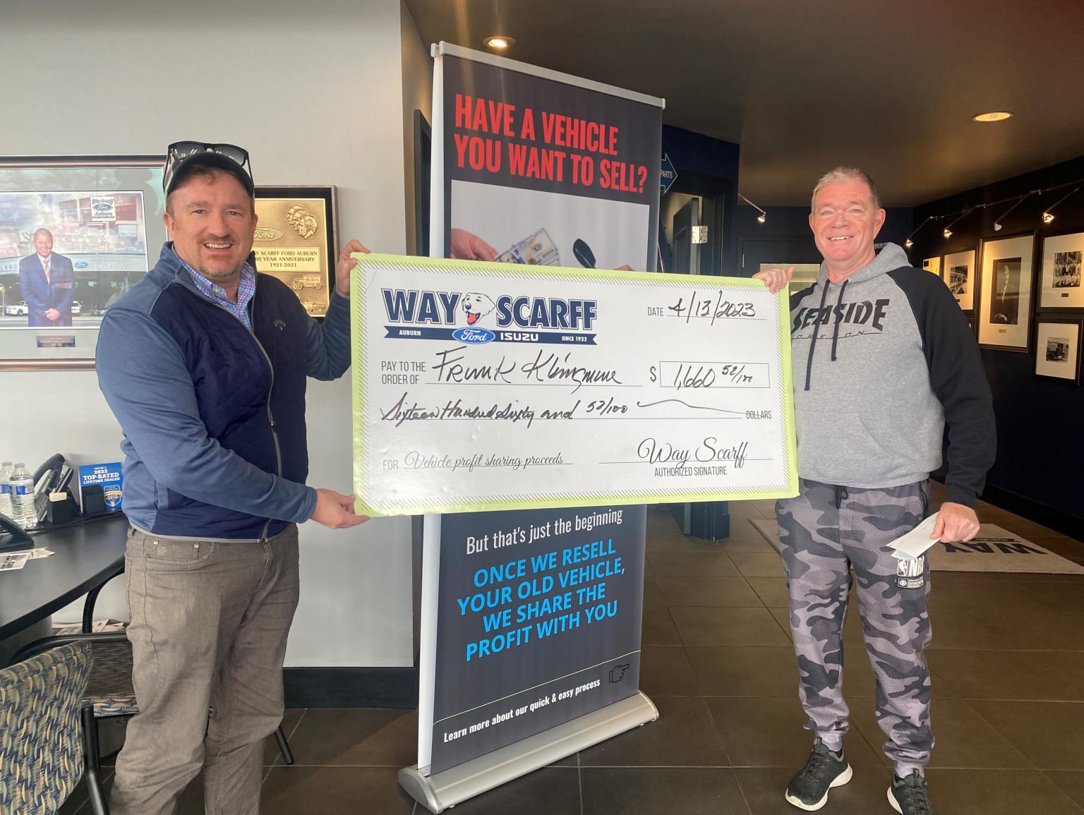 Two men are standing in front of a sign that says "Have a vehicle you want to sell" and holding a large check made out to "Way of the Scarf" for an undisclosed amount.