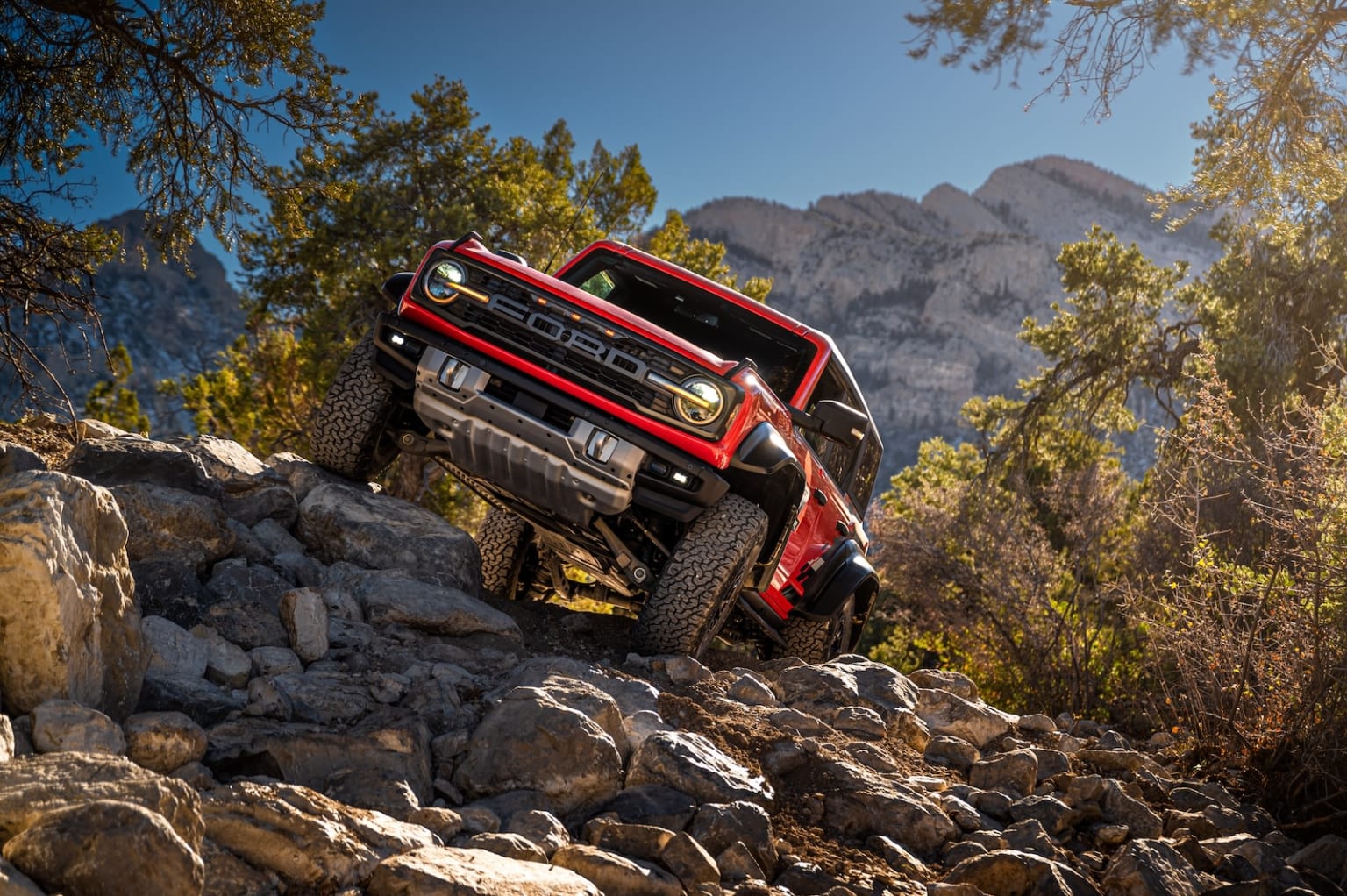 a red truck is driving on a rocky trail with mountains in the backgrouund and trees in the backgrouund