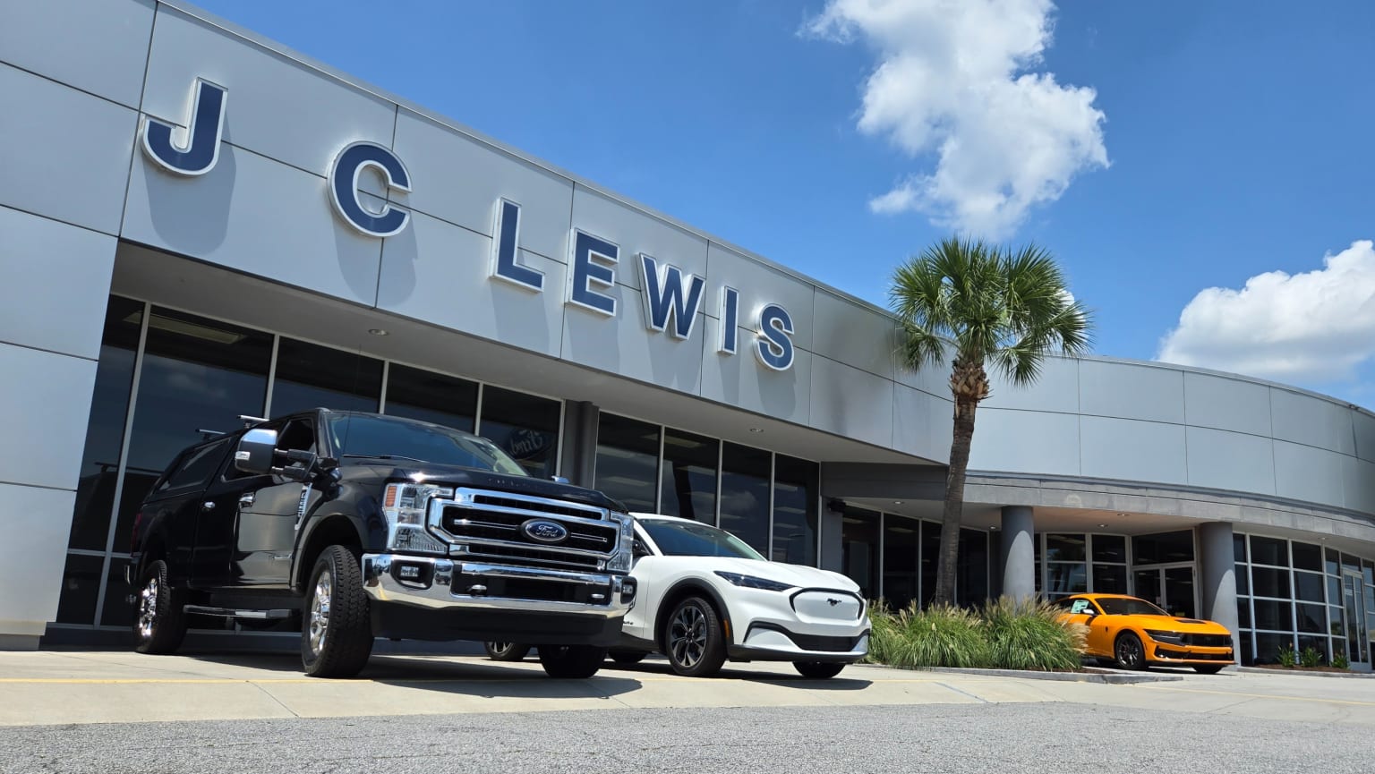 Black truck and white SUV parked in front of a modern car dealership with a bright yellow car visible to the right, under a clear blue sky with fluffy clouds.