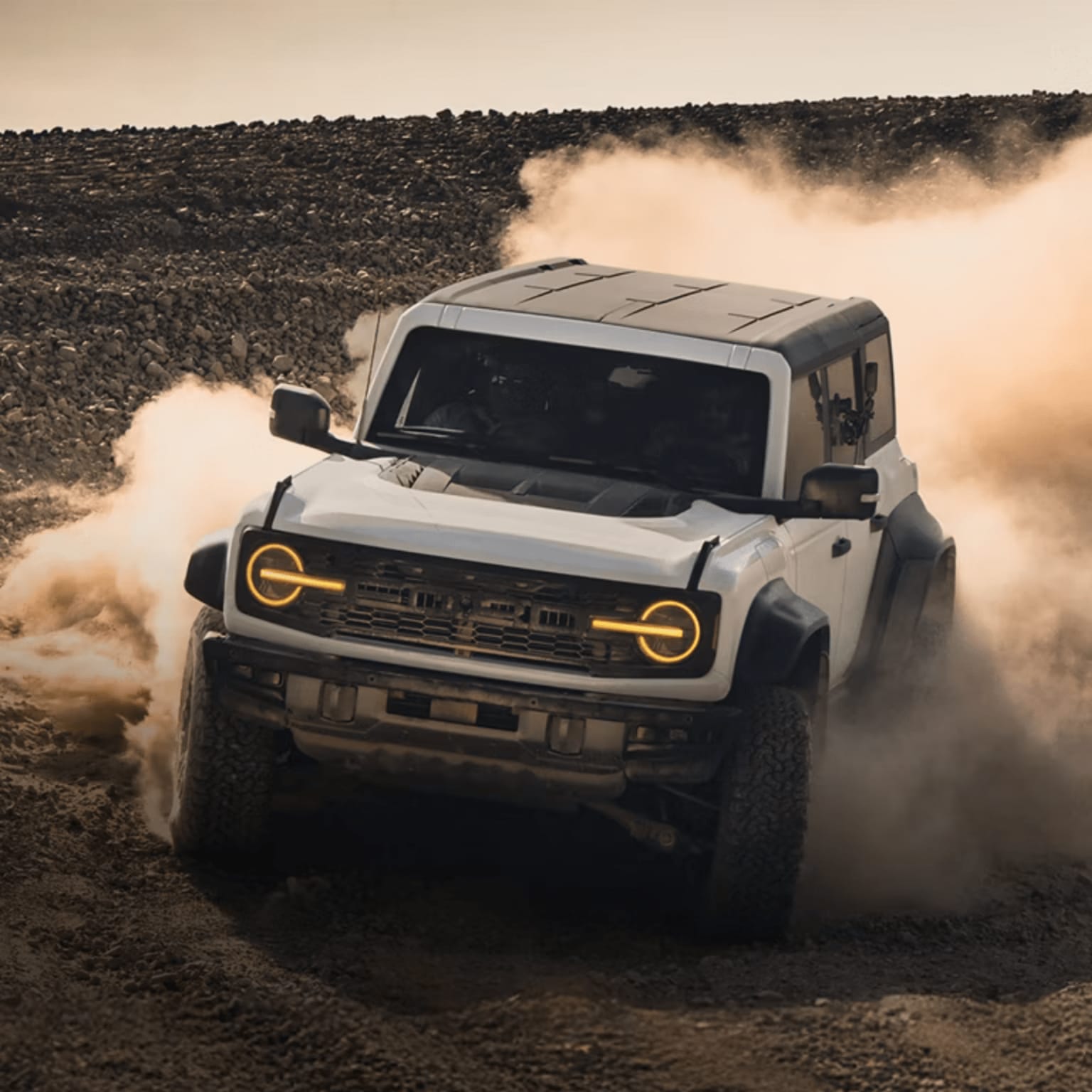 A white SUV kicks up a cloud of dust as it drives on a rocky, barren landscape.