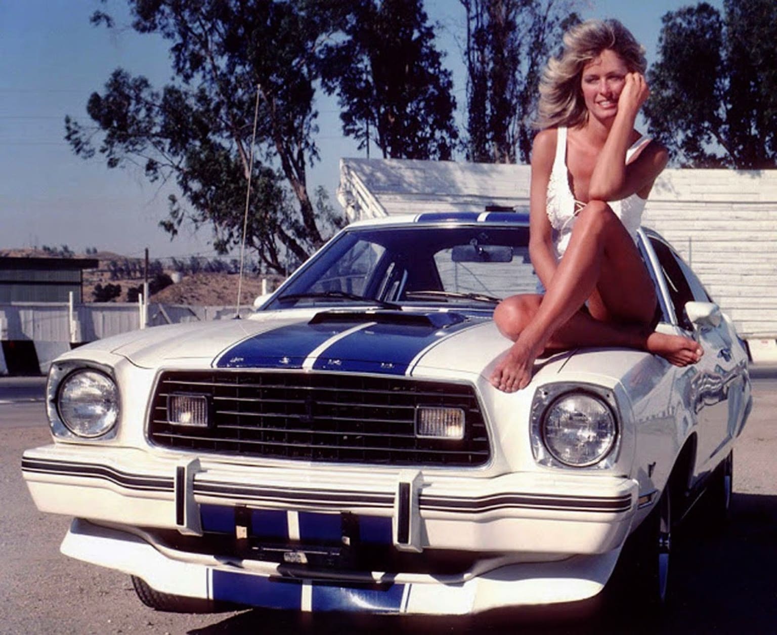 a woman sitting on the hood of a white mustang with a blue and white stripe on the front of it
