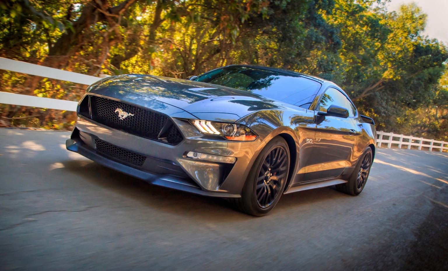 a silver mustang driving down a road next to a white fence and a white fence with trees in the background