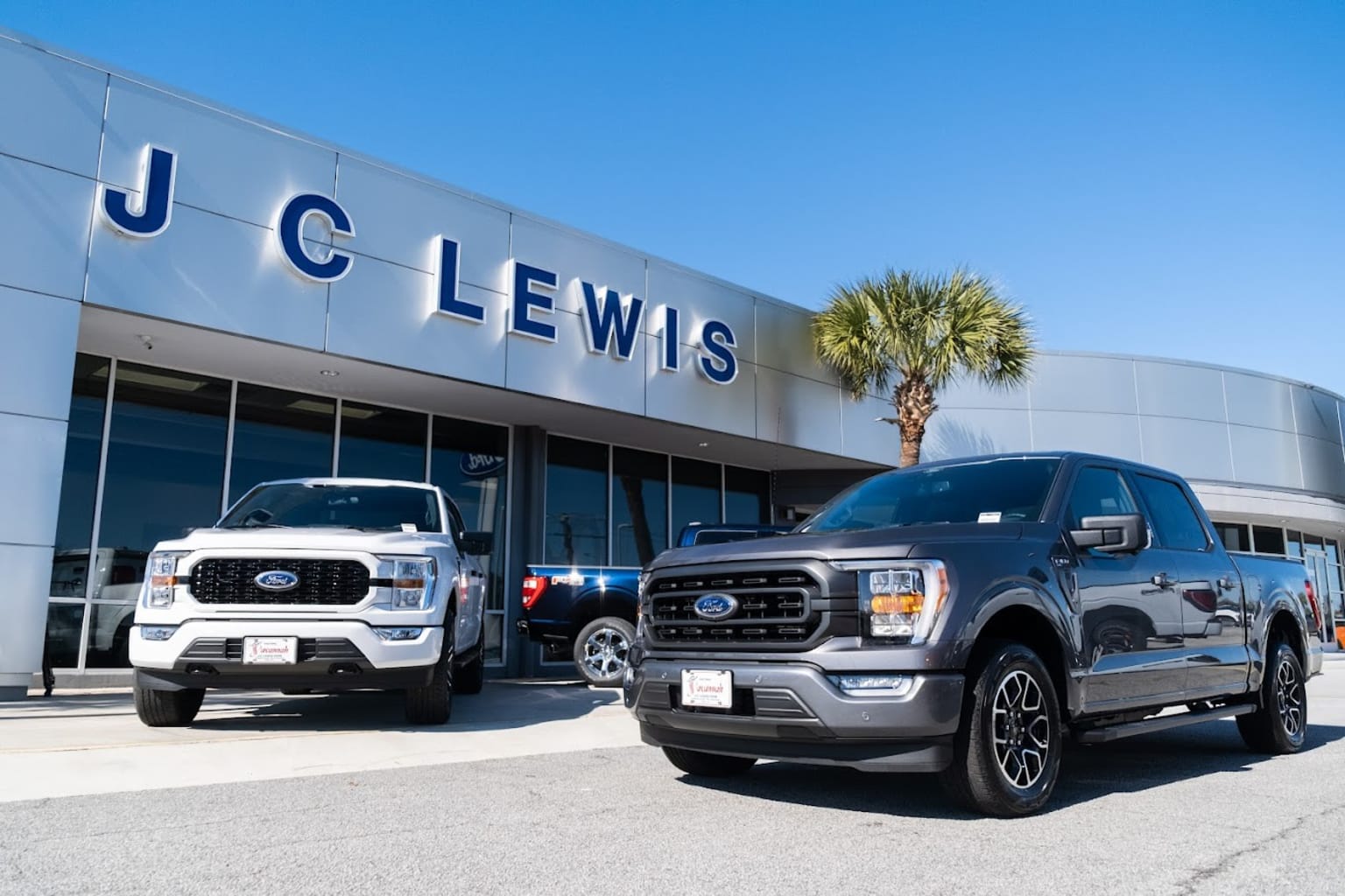 two trucks are parked in front of a dealership with a palm tree in the foreground and a blue sky in the background