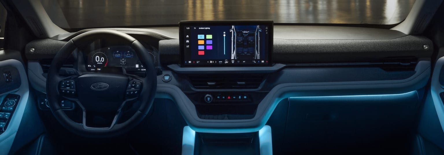 The image shows the interior of a modern vehicle with a large touchscreen display in the center console displaying various controls and information. The steering wheel and dashboard are visible in the foreground, indicating this is the driver's perspective.
