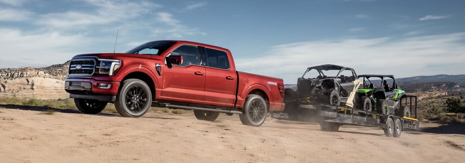 The image shows a red pickup truck towing a trailer with off-road vehicles on a dusty, sandy road against a cloudy sky background.