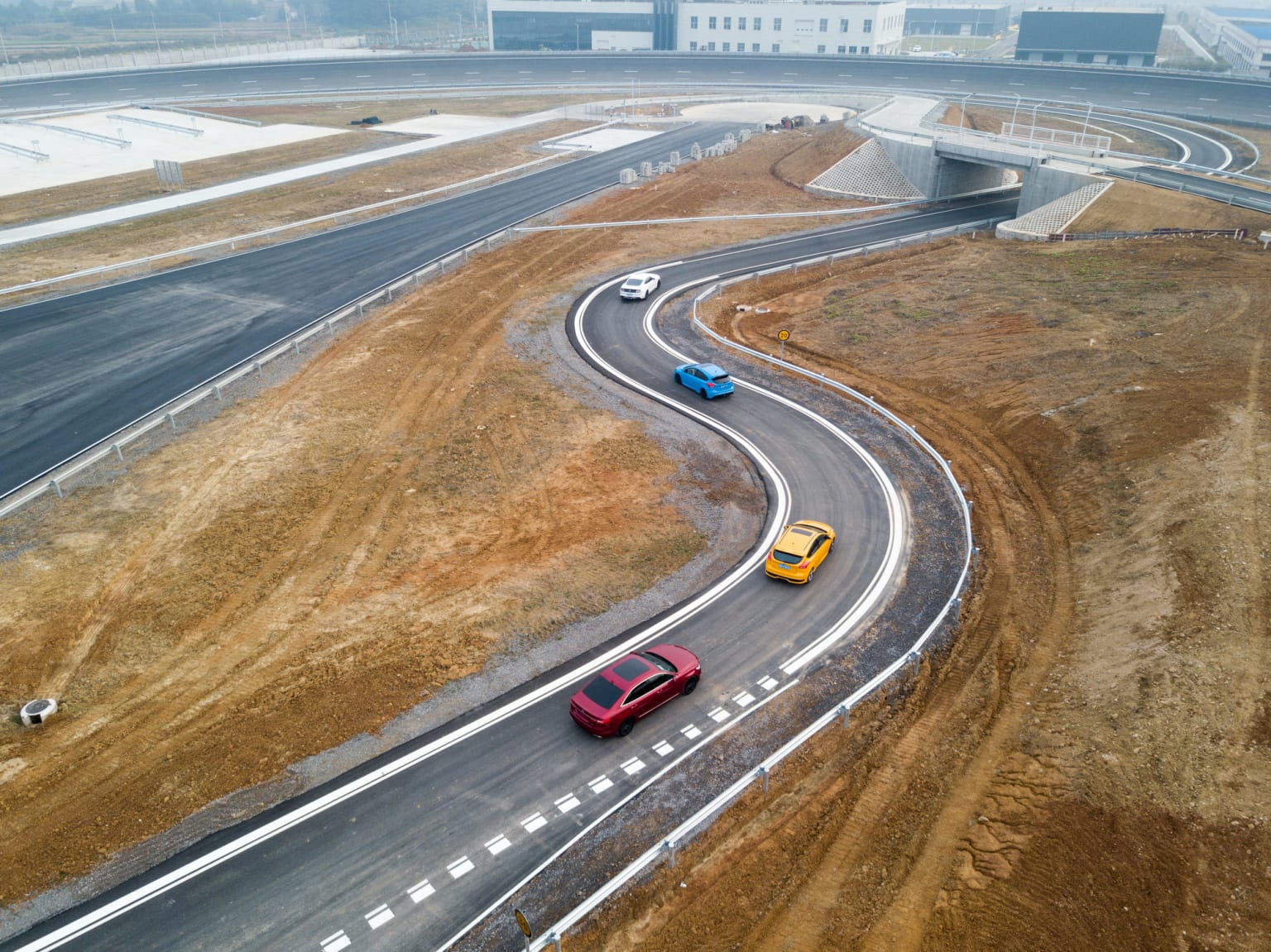 two cars driving down a winding road in the middle of a dry grass and dirt area with a bridge in the background
