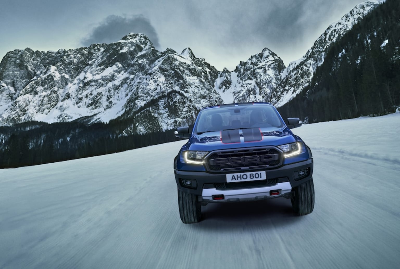 a blue truck driving down a snow covered road next to a large mountain covered with snow covered mountains in the distance