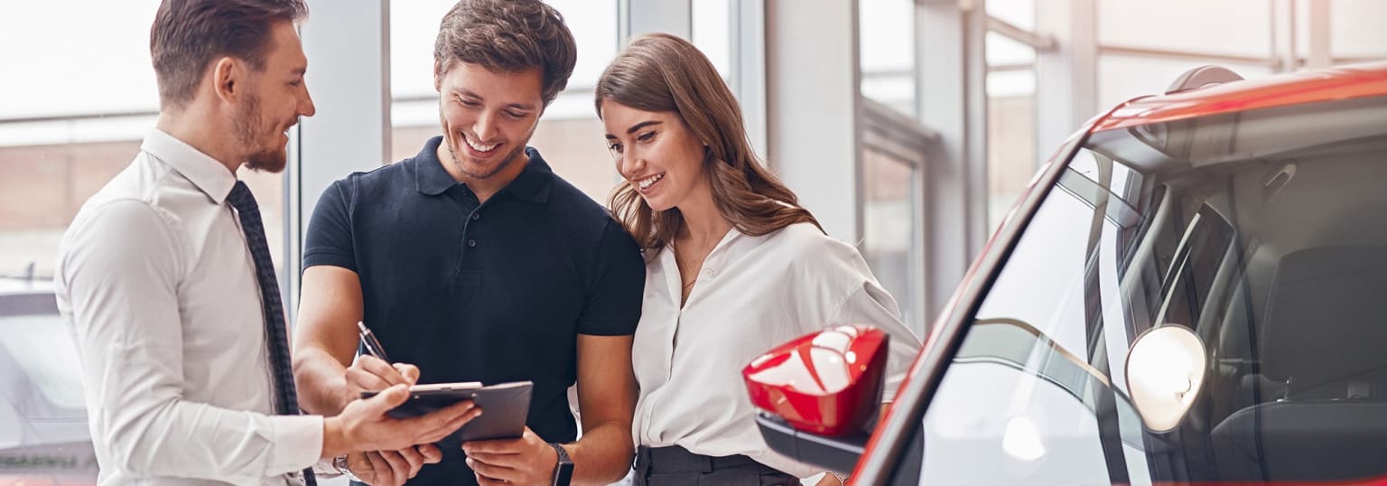 A salesperson showing information on a tablet to a couple in front of a car.