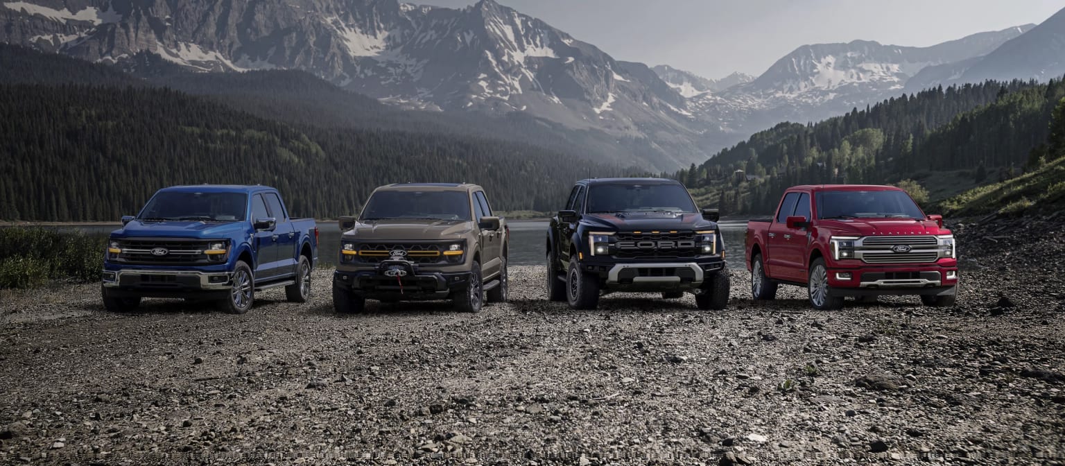 Four pickup trucks in various colors parked on a dirt road with a majestic mountain range in the background.