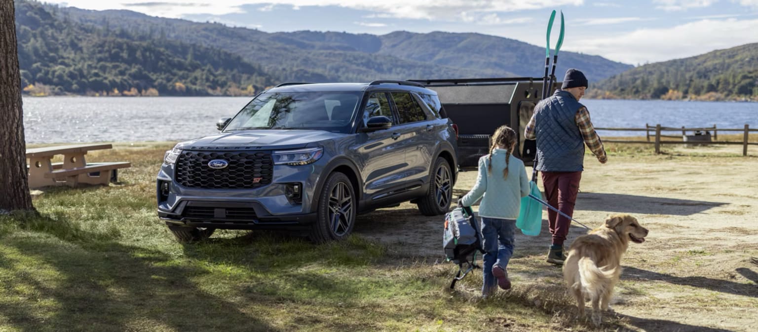 SUV parked on grass near lake with person and dog walking on shore, surrounded by mountainous landscape.