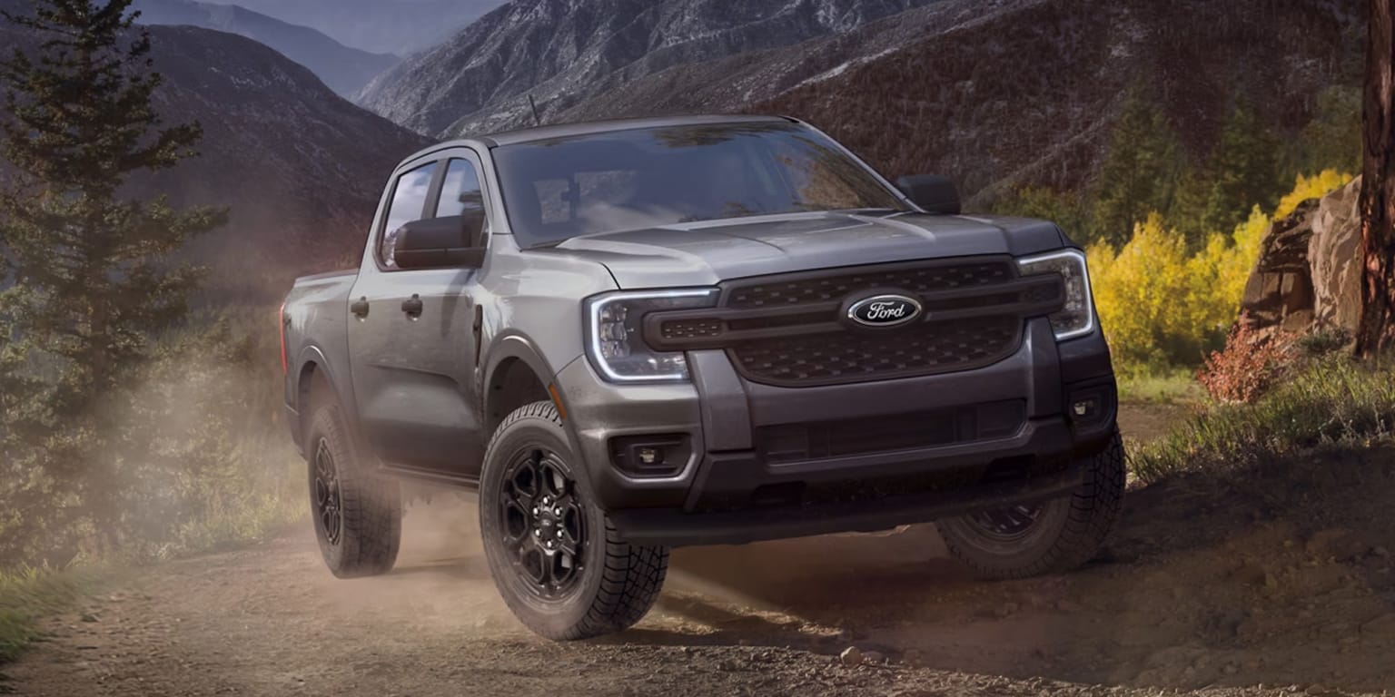 An off-road pickup truck driving along a dusty mountain trail with forested slopes in the background.