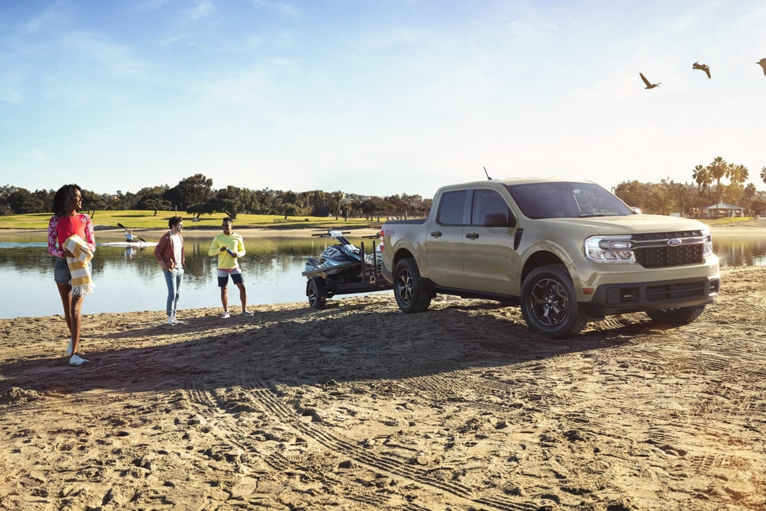 A group of people near a large pickup truck on a sandy beach, with a body of water and trees in the background.