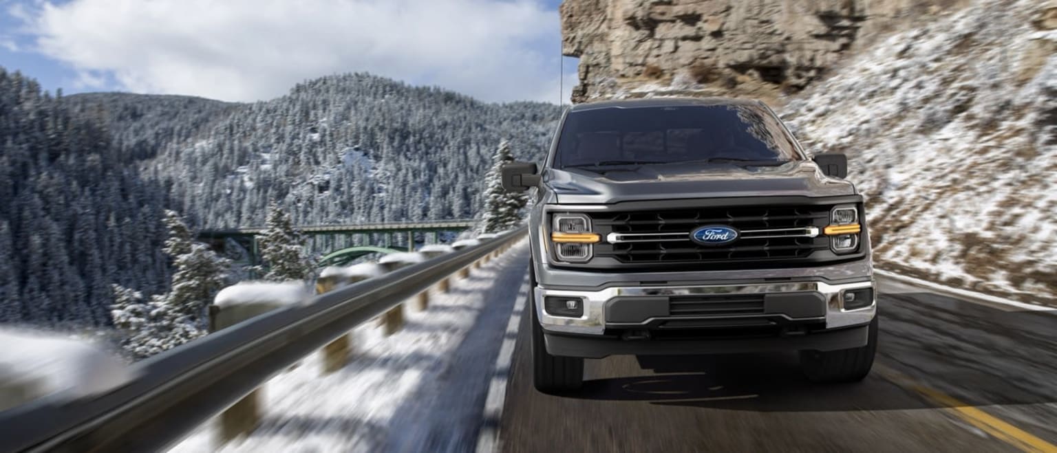A rugged pickup truck navigates a winding mountain road surrounded by snow-capped peaks and dense forests in the background.