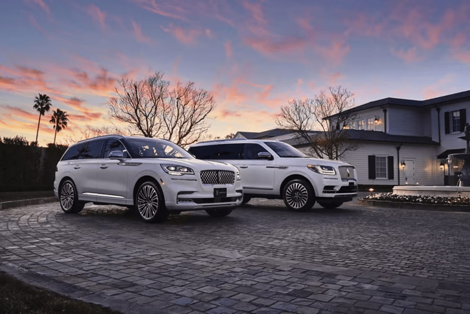 Two white luxury cars are parked side by side in a driveway in front of a house, with a potted plant nearby.