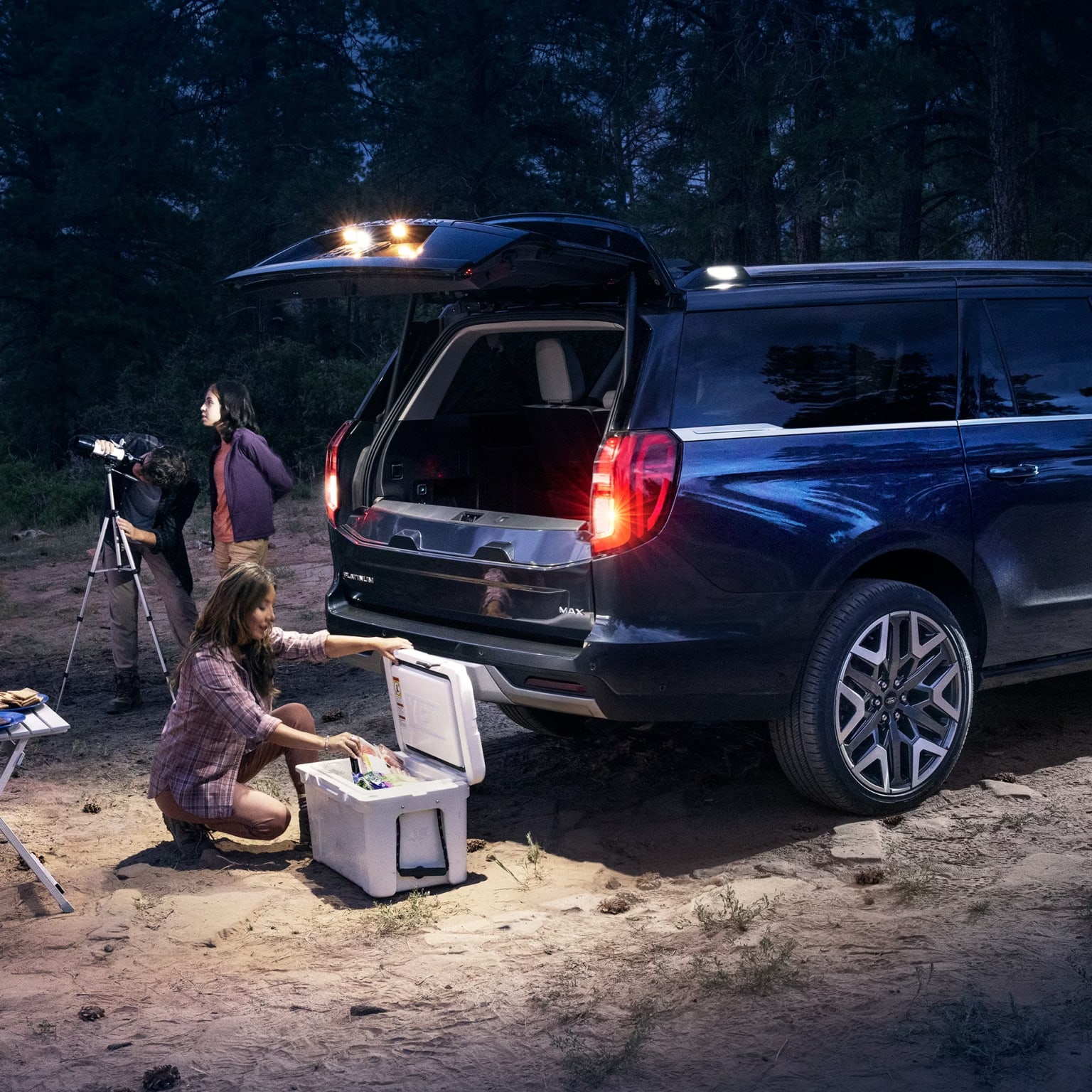 A person sitting on the ground in front of a large SUV with rear hatch open, surrounded by camping equipment and a tripod in a wooded, nighttime setting.