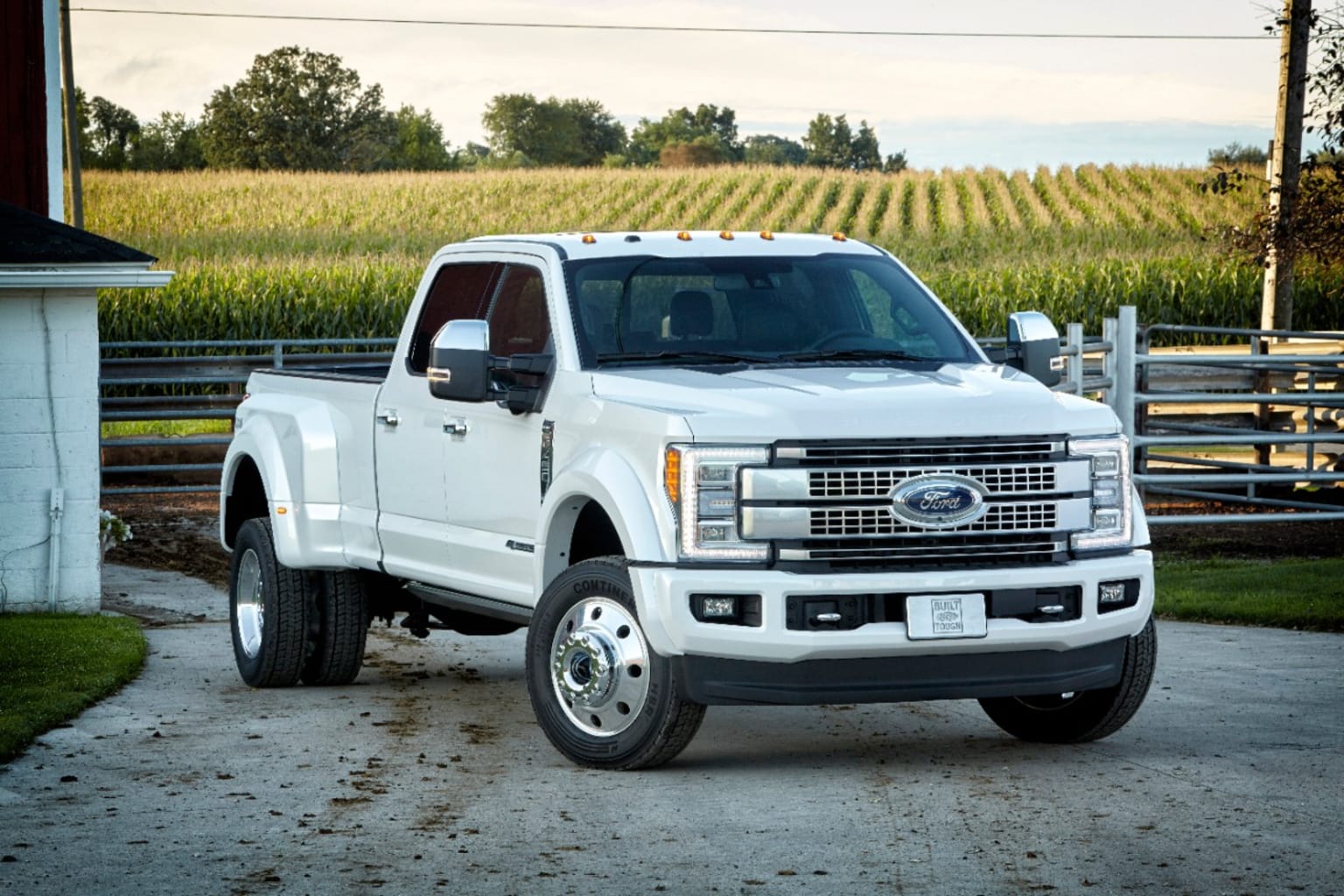 a white pickup truck parked in front of a farm with a cornfield in the background and a barn in the foreground