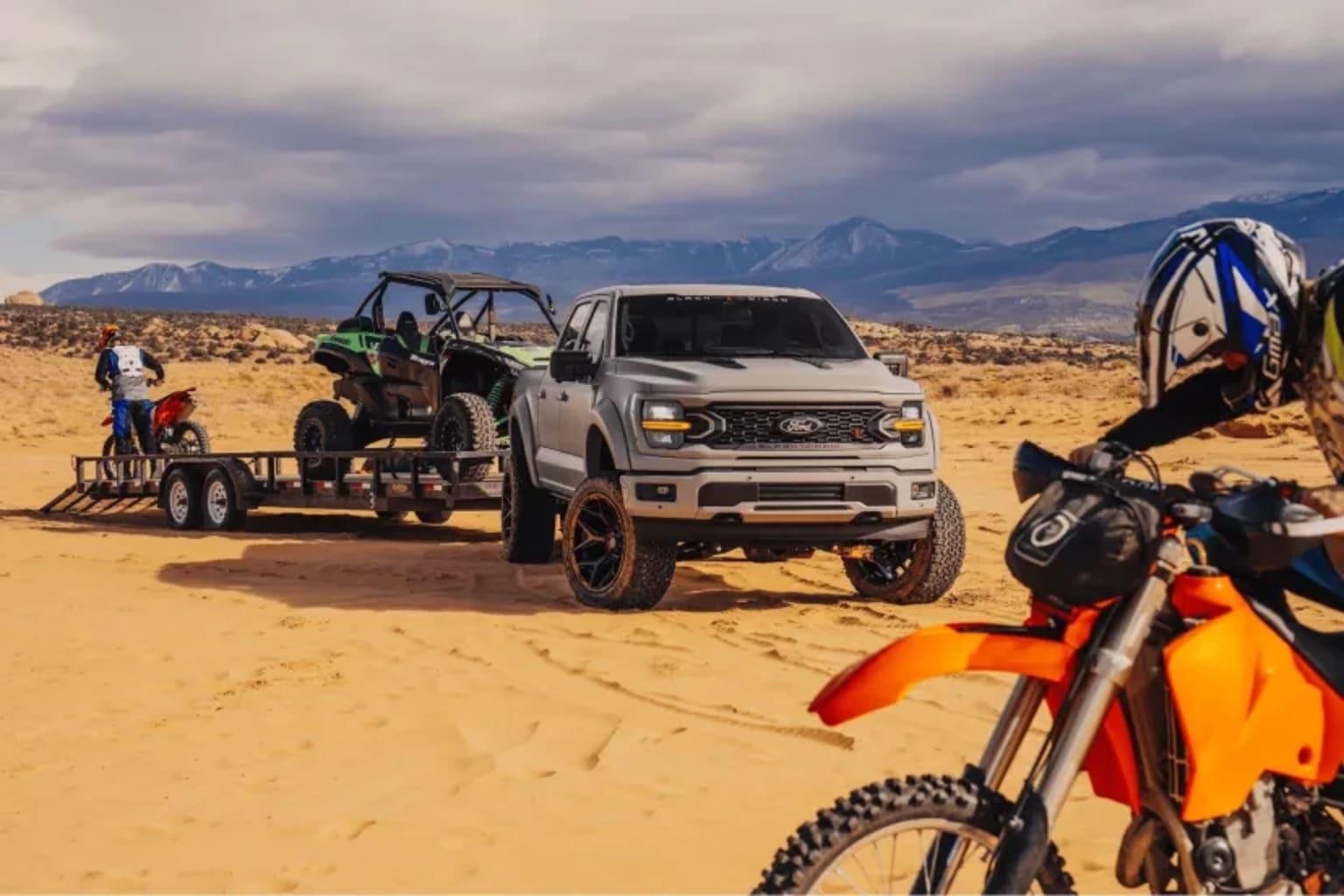 Grey pickup truck towing trailer with side-by-side and dirt bike in desert landscape.